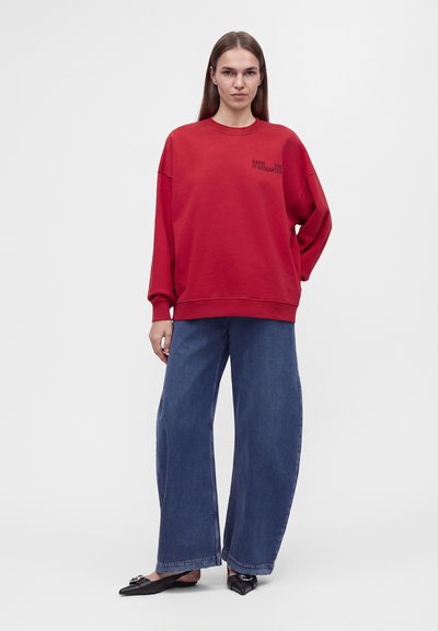 Jeune femme portant un sweat-shirt rouge ample, un jean large bleu et des ballerines noires pointues, debout devant un fond blanc uni.