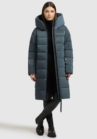 Cappotto lungo imbottito in verde scuro, finitura lucida, con un grande cappuccio, cerniera frontale e polsini a coste. Abbinato a pantaloni neri aderenti e stivali.