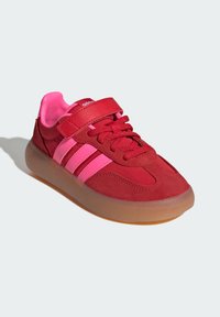 Röda och rosa sneakers med kardborreband, tre rosa ränder på sidan, röda snören och genomskinlig gummisula på vit bakgrund.