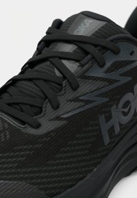 Κοντινό πλάνο ενός μαύρου αθλητικού παπουτσιού με μαύρα κορδόνια, υφασμάτινη υφή και το όνομα της μάρκας «Hoka» στο πλάι.