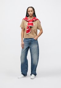 Μπεζ κοντομάνικη μπλούζα με το κείμενο "HUMAN", συνδυασμένη με ανοιχτό μπλε τζιν παντελόνι με φαρδιά μπατζάκια. Πουλόβερ με κόκκινες και λευκές ρίγες δεμένο γύρω από τους ώμους. Λευκά αθλητικά παπούτσια.