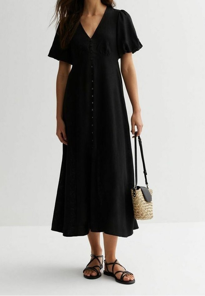 Femme portant une robe midi noire à boutons sur le devant avec des manches bouffantes, tenant un sac seau tissé, et portant des sandales plates noires à brides.