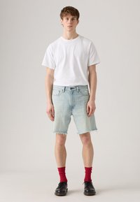 Shorts en denim bleu clair avec ourlet effiloché, associés à un t-shirt blanc uni et des chaussettes rouges, portés avec des chaussures noires.