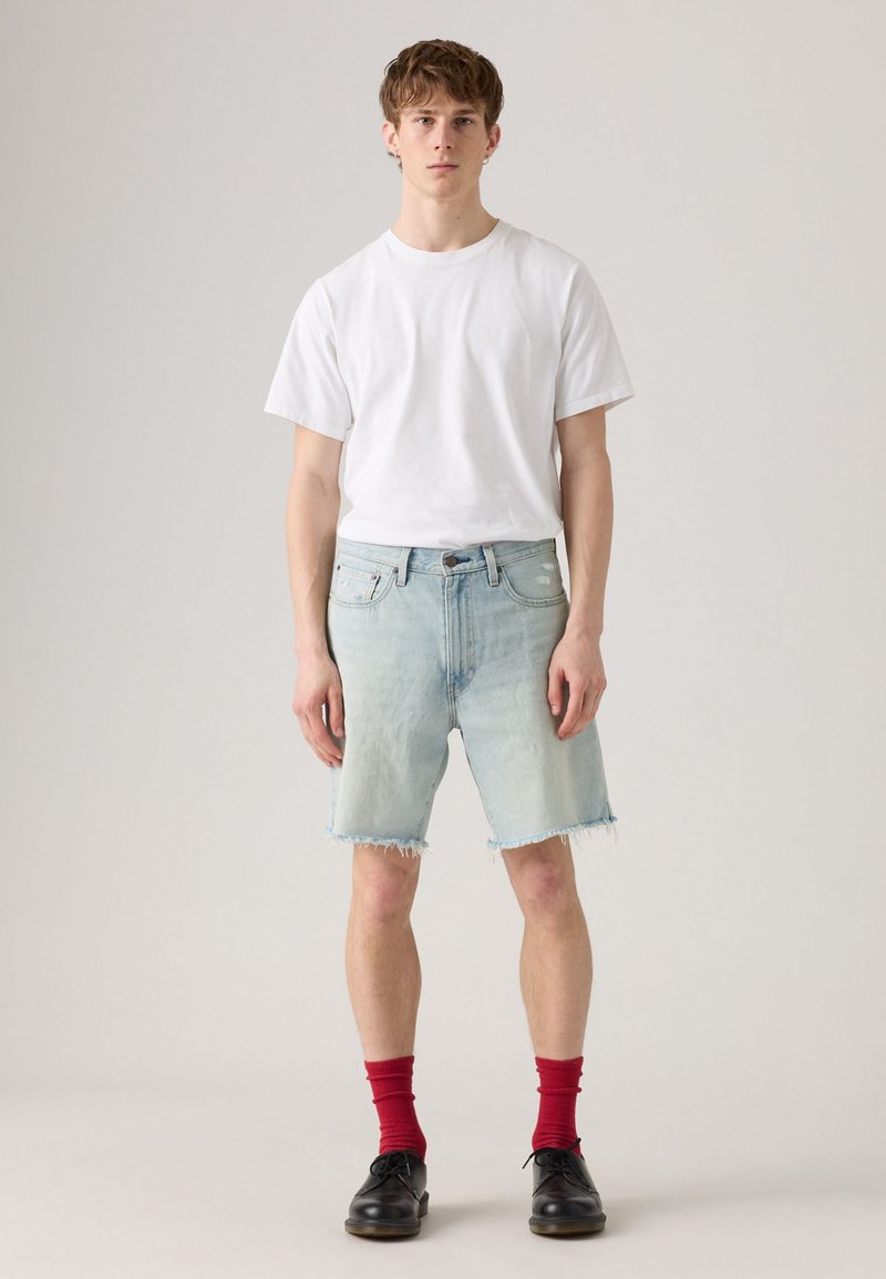 Helle blaue Jeansshorts mit ausgefranstem Saum, kombiniert mit einem schlichten weißen T-Shirt und roten Socken, getragen mit schwarzen Schuhen.