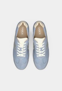 Ανοιχτό μπλε αθλητικά παπούτσια με απαλό υλικό, με λευκά κορδόνια και κρεμ λεπτομέρειες. Χρυσές ρίγες διακοσμούν τις πλευρές. Το όνομα της μάρκας είναι ορατό στο εσωτερικό.