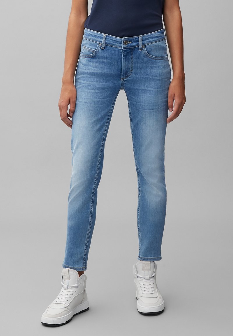Osoba oblečená v světlemodrých slim-fit džínách a bílých vysokých teniskách, stojící na jednotném šedém pozadí.