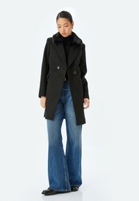 Sort dobbeltbrystet frakke med krave i kunstpels, kombinert med bredbenede blå jeans. Frakken har store knapper og en skræddersyet pasform.