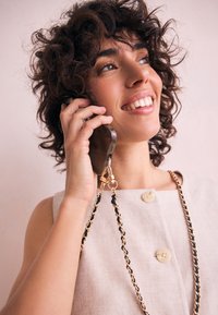 Viedtālrunis ar caurspīdīgu maciņu, pievienots zelta un melnas krāsas ķēdītei. Fona krāsojumā ir maigs rozā tonis. Elegantā, minimālistiskā dizainā.