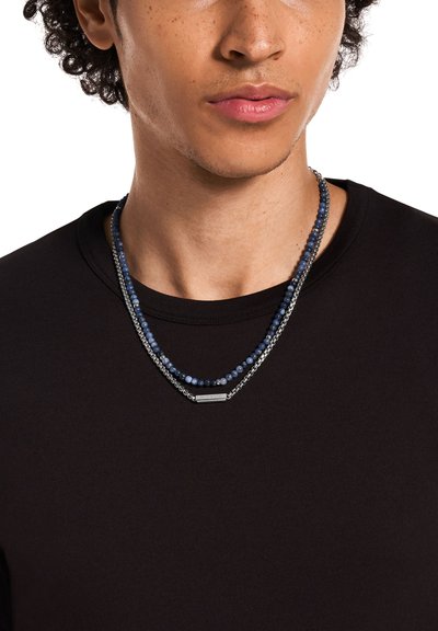 Collier superposé avec des accents de perles bleues et un maillon en métal ton argenté. Porté sur une chemise noire, présentant une texture lisse.