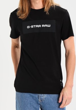 Mężczyzna w czarnej koszulce G-STAR RAW i niebieskich dżinsach, stoi z jedną ręką swobodnie opartą wzdłuż ciała.
