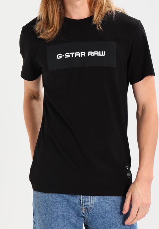 Homme portant un t-shirt noir G-STAR RAW et un jean bleu, debout avec une main détendue sur le côté.