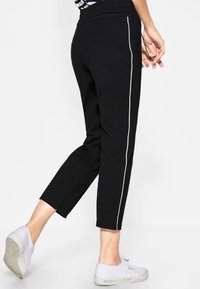 Czarne spodnie typu crop z elastycznym pasem, z białymi bocznymi paskami i gładką, śliską fakturą. Model ma na sobie białe trampki.