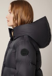 Czarna ocieplana kurtka z dużym kapturem, wykonana z teksturowanej tkaniny, z pikowanym wzorem i okrągłą łatą marki na rękawie.