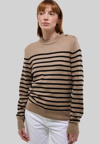 Femme portant un pull beige et noir à rayures avec des boutons sur l'épaule, associé à un pantalon blanc, sur un fond gris uni.