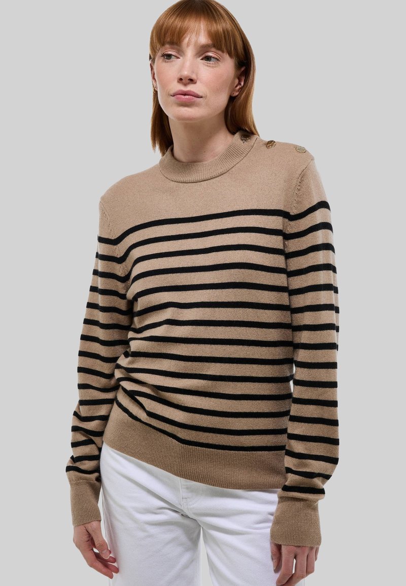 Femme portant un pull beige et noir à rayures avec des boutons sur l'épaule, associé à un pantalon blanc, sur un fond gris uni.