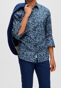 Blå mönstrad skjorta med blommönster, lång ärm och knappstängning, bärs med en marinblå blazer över axeln och marinblå byxor.