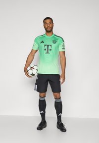 Jersey jalkapallolle, jossa on vihreä gradientti, mustat akcentit, logo ja sponsoriteksti. Mustat shortsit, mustat sukat valkoisilla raidoilla ja jalkapallo.