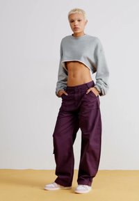 Sweatshirt gris coupé avec des manches longues, associé à un pantalon cargo ample de couleur violet profond. Le modèle porte des baskets blanches.