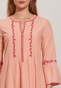 Femme portant un chemisier rose pâle avec des plis verticaux et une broderie florale rose foncé sur le devant et les manches, avec un col à volants.