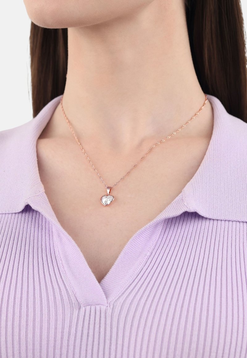 Collana a ciondolo a forma di cuore in oro rosa con una pietra trasparente, su una catena attorcigliata, indossata sopra un top viola a coste con colletto.