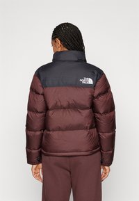 Doudoune bordeaux et marine avec un col montant, présentant des panneaux matelassés et un logo au dos. Texture douce et poignets élastiques.