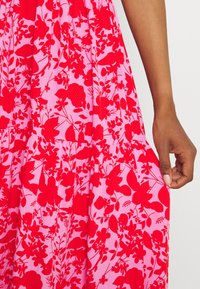 Robe à imprimé floral rouge sur tissu rose clair, caractérisée par une taille froncée et des détails texturés. Main tenant le tissu sur le côté.