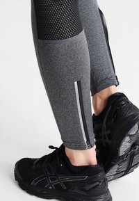 Harmaat urheiluleggingsit, joissa on verkkoelementtejä, sivuvetoketjut heijastavilla yksityiskohdilla, yhdistettynä mustiin juoksukenkiin.