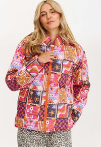 Kurtka pikowana z żywym, wielopatternowym wzorem zawierającym motywy kwiatowe, oczu oraz geometryczne. Pomarańczowy zamek, wysoki kołnierz i kontrastowe mankiety.