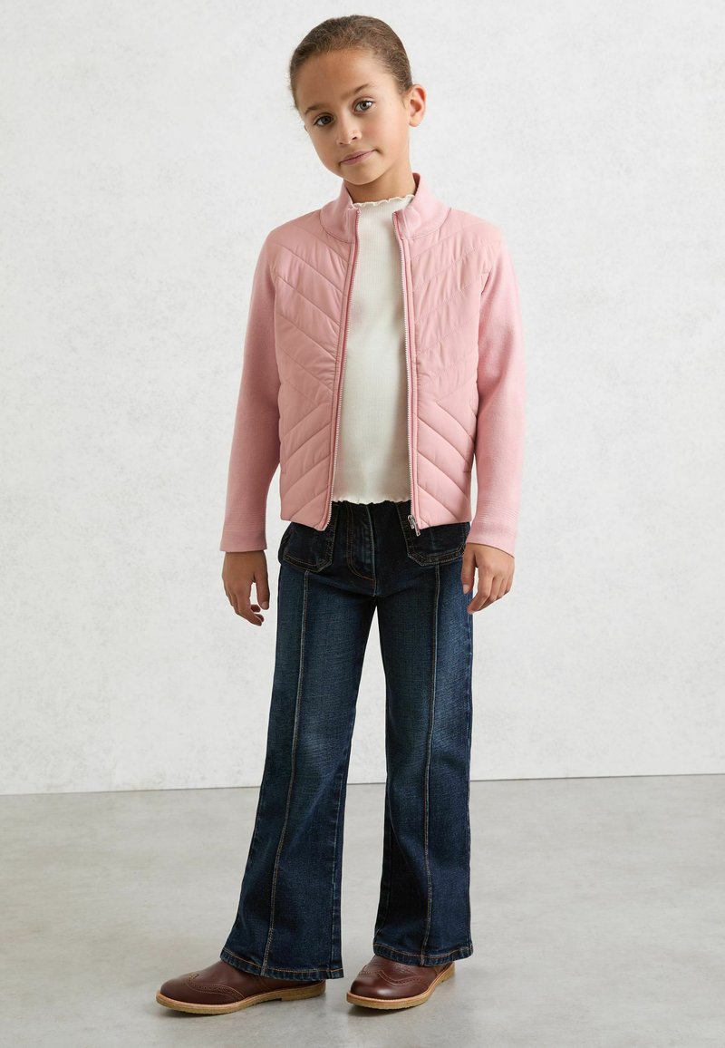 Lyserød quiltet jakke med høj krave, kombineret med en hvid tekstureret top og mørkeblå vide jeans. Brune læderstøvler fuldender outfittet.