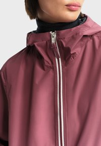 Žena u ljubičastoj kaputu s kapuljačom, zatvorenoj do kraja, preko crnog rolerca, prikazana od vrata do gornjeg dijela prsnog koša.