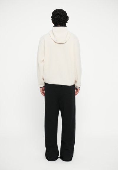Helle beige Kapuzenjacke von hinten, mit einer Kordelzugkapuze und gerippten Bündchen, kombiniert mit schwarzen, weit geschnittenen Hosen. Glatte Textur.