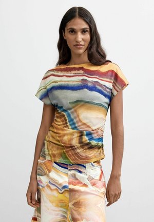 Mujer con cabello largo y oscuro que lleva una blusa de manga corta con un patrón abstracto y colorido, y una falda a juego, sobre un fondo blanco liso.