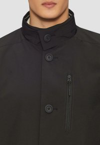 Cappotto nero con colletto alto, dotato di due grandi bottoni, una tasca sul petto con zip e una texture liscia e opaca.
