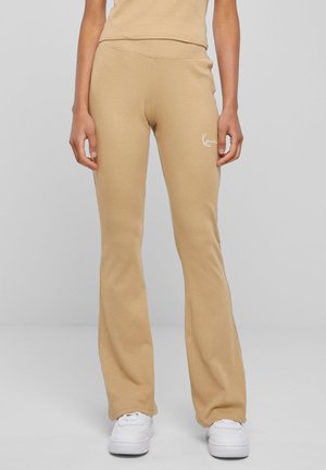 Beige, utsvingte bukser laget av ribbet stoff, med høy midje og en liten hvit logo på hoften. Brukt med hvite sneakers.