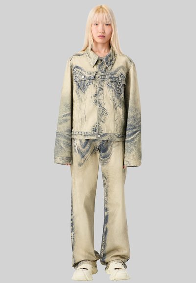 Beige denimjakke og bukser med blå teksturerede mønstre, løs pasform. Model der bærer hvide sneakers, står foran en grå baggrund.