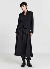 Blazer nero con risvolti arrotondati e tre bottoni dorati, abbinato a una gonna nera fluida e stivali neri al ginocchio. Tessuto liscio.