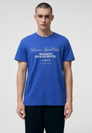 Młody mężczyzna z krótkimi włosami, ubrany w niebieski T-shirt z białym napisem "Diverse Sport Club Monaco Grand Hotel" oraz pięcioma gwiazdkami, stojący zwrócony przodem.