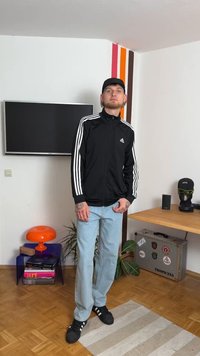 Svart zip-up jacka med vita ränder, ljusblå jeans och svarta sneakers. Tillbehör inkluderar en keps och synliga tatueringar på händerna.