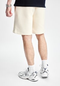Beige bomullsblandede shorts med en avslappet passform, som har en glatt tekstur og kantede kanter, kombinert med hvite sportsokker og grå sneakers.