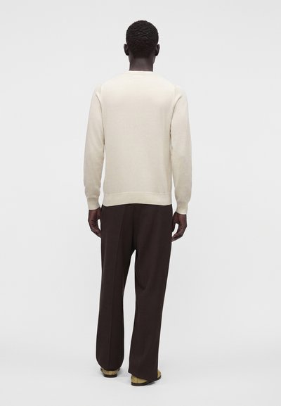 Homme aux cheveux courts et bouclés portant un pull beige à manches longues, un pantalon large sombre et des chaussures marron, debout de dos sur un fond uni.