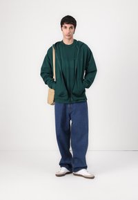 Grønn zip-up hettegenser over en matchende T-skjorte, parret med blå løse jeans og hvite sneakers. En brun skulderveske bæres på den ene siden.