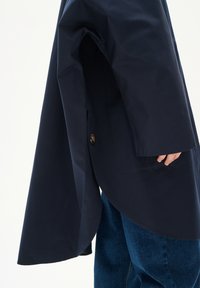 Morska dežna jopica s ukrivljenim robom, izdelana iz vodoodpornega materiala. Ima gumbno zapiranje in široke rokave. V kompletu z jeans hlačami.