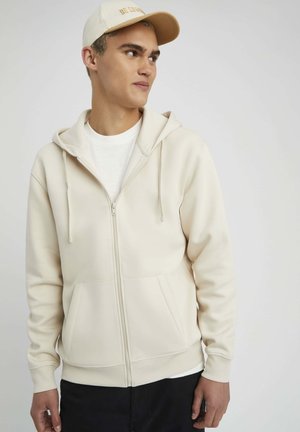Haut à capuche beige clair zippé en tissu doux avec une poche kangourou, une capuche à cordon et un t-shirt blanc en dessous. Design simple et moderne.