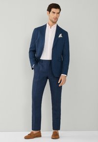 Completo in lino blu navy con vestibilità su misura, abbinato a una camicia button-up rosa chiaro. Mocassini marroni e un fazzoletto bianco completano l'outfit.