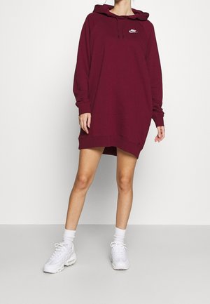 Vrouw draagt een bordeauxrode Nike hoodie jurk met lange mouwen en witte sneakers, staand tegen een effen lichte achtergrond.