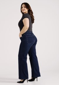 Donkerblauwe high-waisted jeans met uitlopende pijpen, voorkeurszakken, en een donkere, aansluitende shirt, gestyled met zwarte hoge hakken.