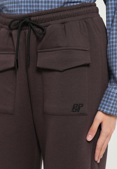 Σκούρες καφέ φόρμες jogger με ελαστική μέση και κορδόνι, δύο μπροστινές τσέπες και μαύρο κεντημένο λογότυπο στο πλάι. Λείος υφή και μαλακό υλικό.