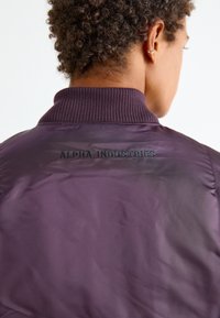 Paarse bomberjack met een geribbelde kraag en de tekst "ALPHA INDUSTRIES" geborduurd in donkere draad op de achterkant. Gladde textuur.