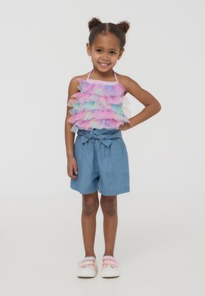 Top in tulle multicolore con strati arricciati, abbinato a pantaloni corti in denim blu e sneakers pastello con strappi in Velcro.