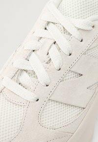 Gros plan sur une basket blanche montrant un tissu texturé, des empiècements en daim, des détails cousus et des lacets tissés épais noués selon un motif de laçage standard.